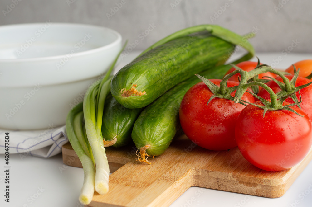 On a light background, a cutting board with tomatoes and cucumbers, green onions, a white plate. The concept of healthy food, vegetarianism, diet. On a light background, a cutting board with tomatoes and cucumbers, green onions, a white plate. The concept of healthy food, vegetarianism, diet.