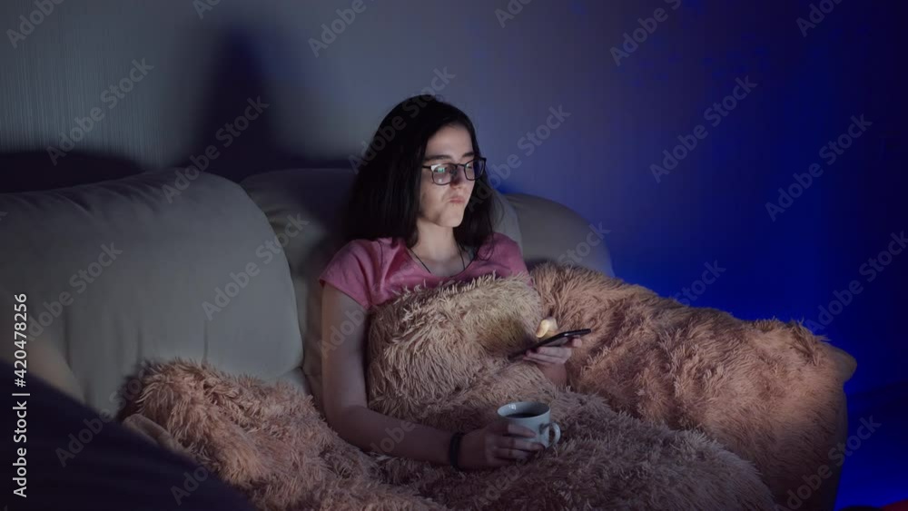 girl with a cup of tea sits on the sofa in the evening and watches tv, glare on glasses