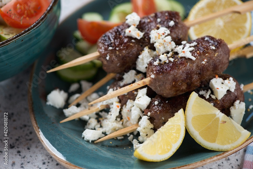 Close-up of roasted greek kofta kebabs with feta cheese, fresh vegetables and lemon on a turquoise plate, selective focus photo