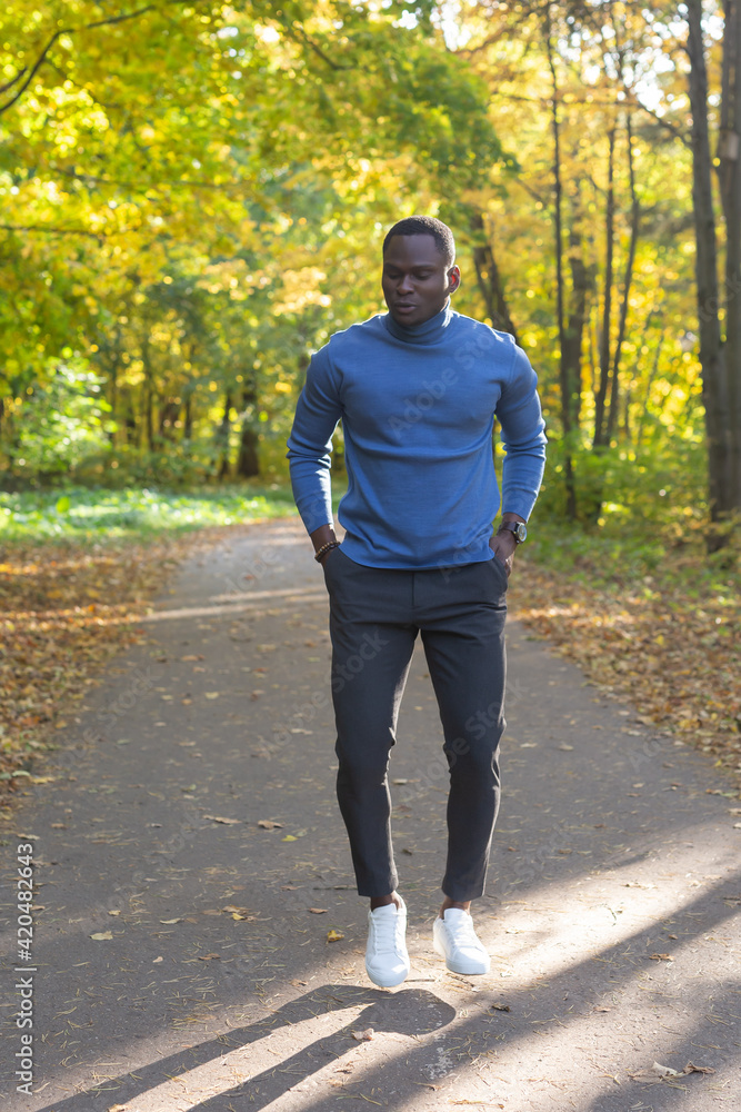 Obraz premium Cheerful african american young man in stylish clothes walks in autumn park on sunny warm autumn day. Concept of weekend and outdoor recreation