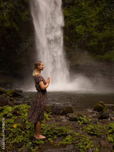 Fototapeta Naklejka Na Ścianę i Meble -  Young Caucasian woman spending time near waterfall. Meditation and relaxation. Hands in namaste mudra. Closed eyes. Travel lifestyle. Woman wearing dress. Nung Nung waterfall in Bali