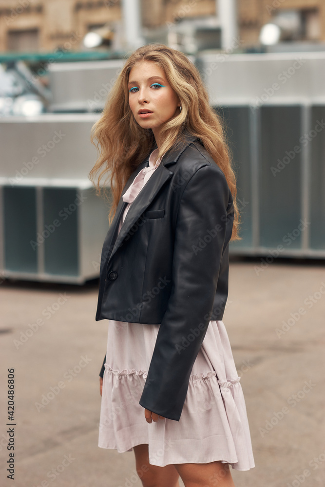 Fototapeta premium Young stylish woman in pink dress, black leather jacket and boots walking at urban location. Female model with long wavy blonde hair outdoor fashion look.