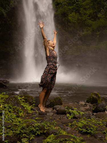 Happy Caucasian woman jumping near the waterfall. Hands in motion movement. Nature and environment concept. Travel lifestyle. Woman wearing dress. Copy space. Nung Nung waterfall in Bali