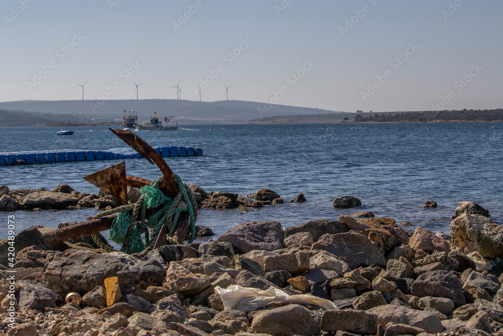 rusted anchor standing between stones on the shore and rope around it. Ship, pier and mediterranean sea in the background with a wide depth of field