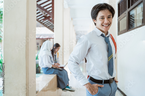 a high school boy smiles at the camera wearing a school bag in the background of a building corridor