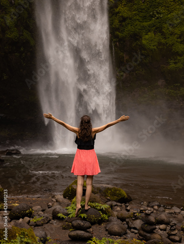 Traveler woman wearing pink dress at waterfall. Excited woman raising arms in front of waterfall. Travel lifestyle. Energy of water. View from back. Copy space. Nung Nung waterfall in Bali