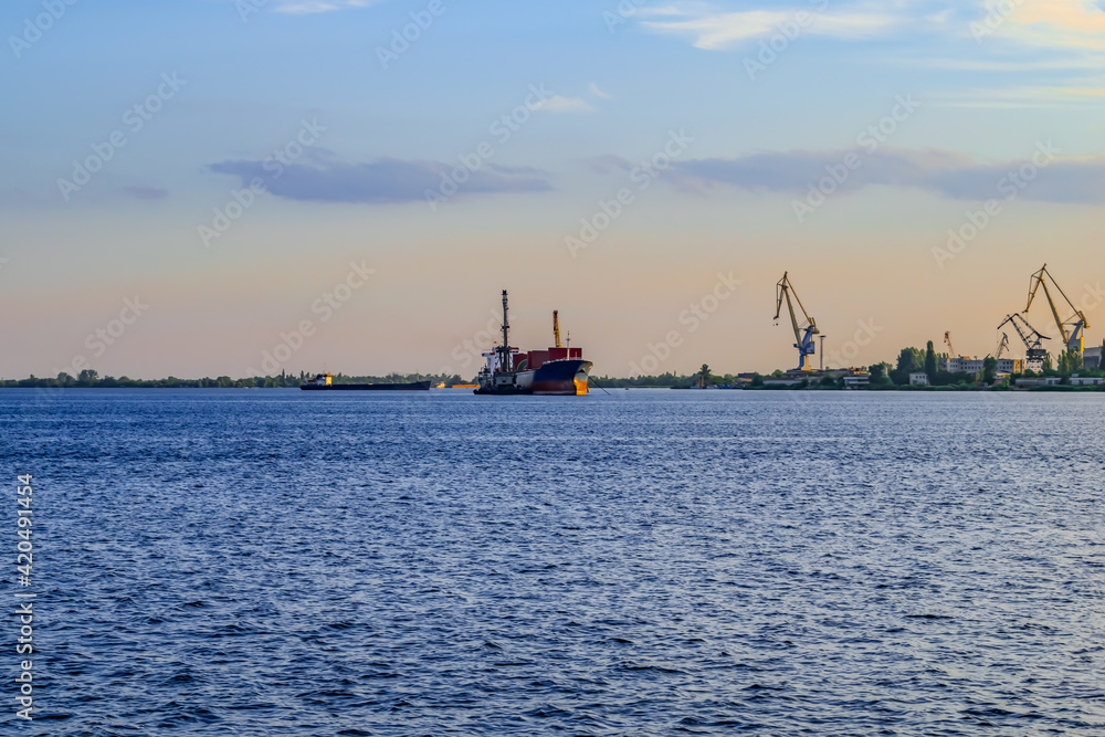 A large cargo ship departs from the Kherson port on the Dnieper river (Ukraine). Evening panorama with watercraft on the blue surface of the water and harbor cranes on the shore