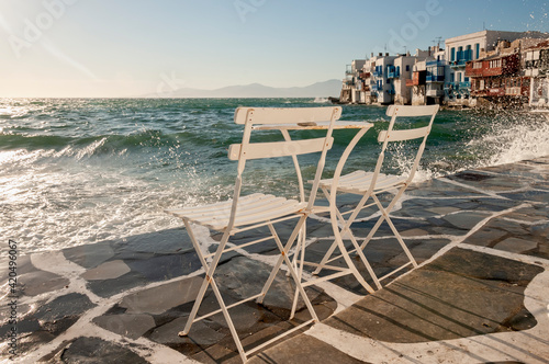 Two empty chairs on the waterfront in the island of Mykonos  Greece. In the background the splash of the waves and the characteristic Greek houses on the coast.