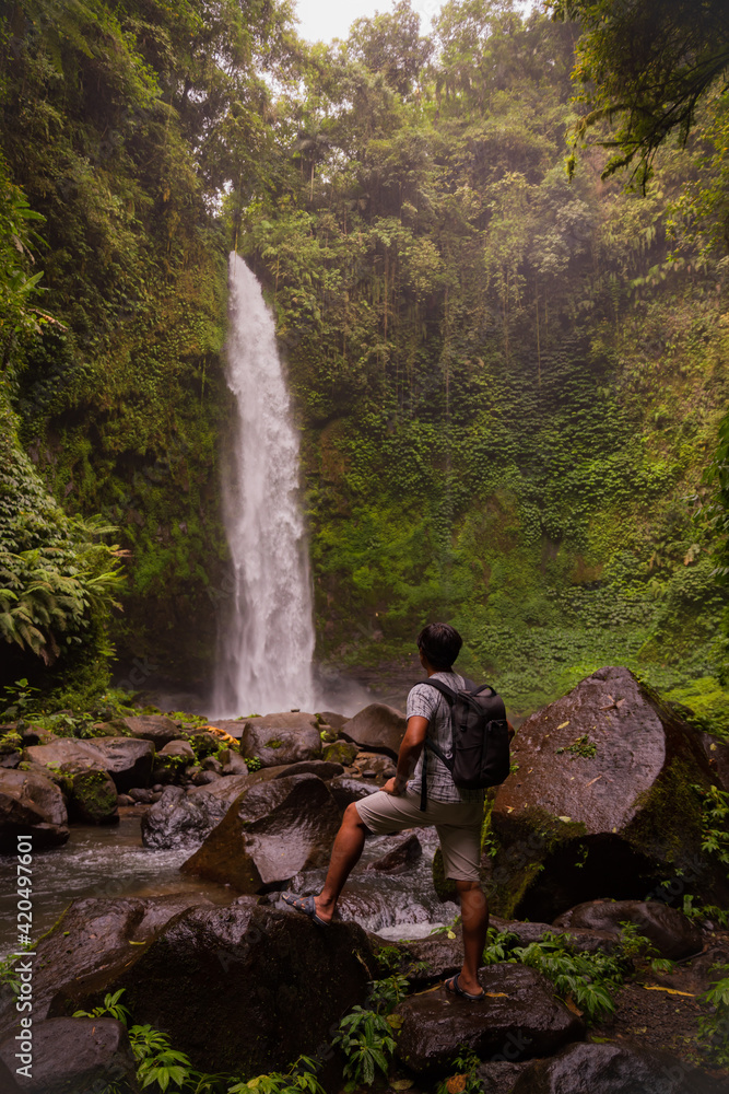 Asian man traveller enjoying waterfall landscape in tropical forest. Man with backpack. Energy of water. Travel lifestyle. View from back. Nung Nung waterfall, Bali