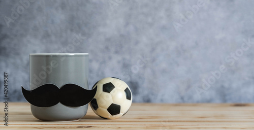 Objects on the table of Happy Fathers day holiday background concept.Black mustache on cup with football on blurred grey backdrop.Sign for the season for dad.mock up, border,banner, copy space.