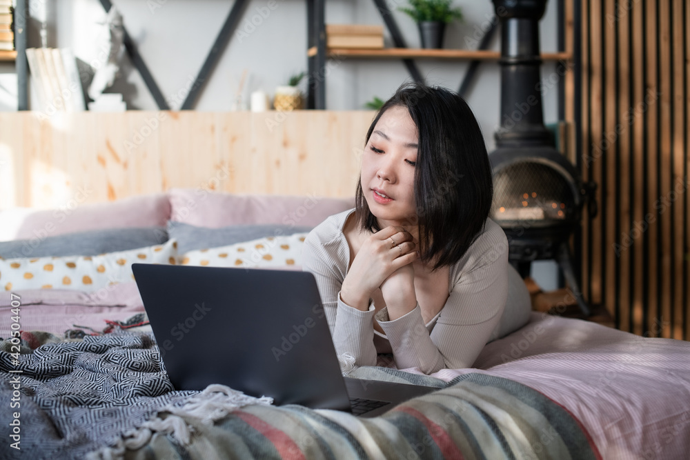 Young Korean woman in home clothes of pastel colors in the bedroom on the bed uses a laptop and relaxes in the interior of the loft and boho. Self isolation. Message and dating in social net. Young Korean woman in home clothes of pastel colors in the bedroom on the bed uses a laptop and relaxes in the interior of the loft and boho. Self isolation. Message and dating in social net.