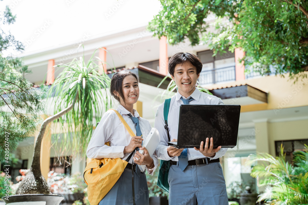 © Odua Images - boy and girl of Indonesian high school students smiling at the camera as they stand holding book, computer laptop, and wearing bag against the background of the school building © Odua Images - boy and girl of Indonesian high school students smiling at the camera as they stand holding book, computer laptop, and wearing bag against the background of the school building