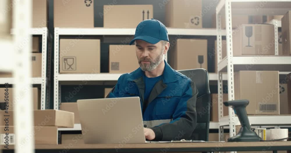 Senior courier sits in warehouse at desk, prepares parcels for shipment and scans boxes by entering data into laptop. Senior courier sits in warehouse at desk, prepares parcels for shipment and scans boxes by entering data into laptop.