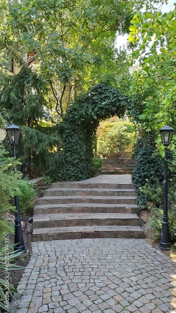 Path through arched gateway to English garden with retro streetlights by sides. Stone paved stairs in park with hedge arch