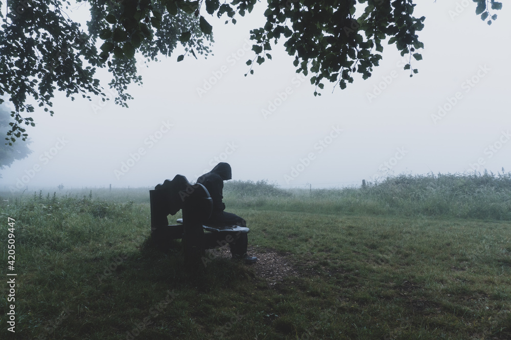 A man sitting on a bench underneath a tree on a moody foggy day A man sitting on a bench underneath a tree on a moody foggy day
