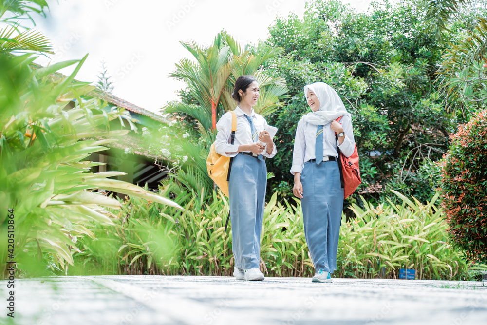 two female high school students wearing school bags while standing and chatting in the school yard with lots of green plants as the background two female high school students wearing school bags while standing and chatting in the school yard with lots of green plants as the background