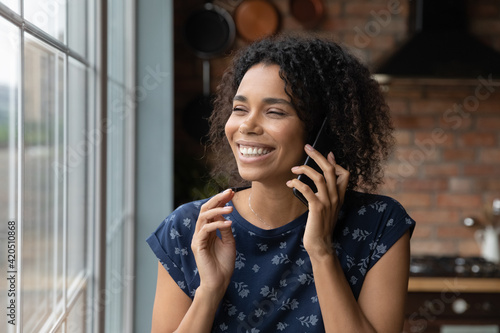 Glad young african ethnic woman talk on telephone call from home stand by window hold device to ear. Happy black housewife caller enjoy phone conversation chat with friend discuss sale share good news