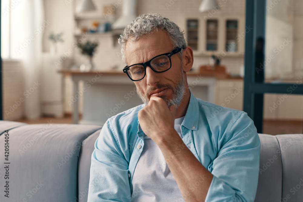 Naklejka premium Relaxed senior man in casual clothing and eyeglasses keeping hand on chin while resting on the sofa at home