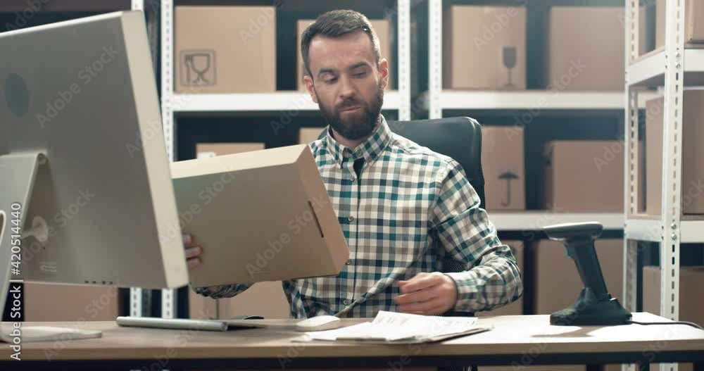 Logistics manager sits at desk in warehouse on background of cardboard boxes, and scanning parcel, works with information on computer. Logistics manager sits at desk in warehouse on background of cardboard boxes, and scanning parcel, works with information on computer.