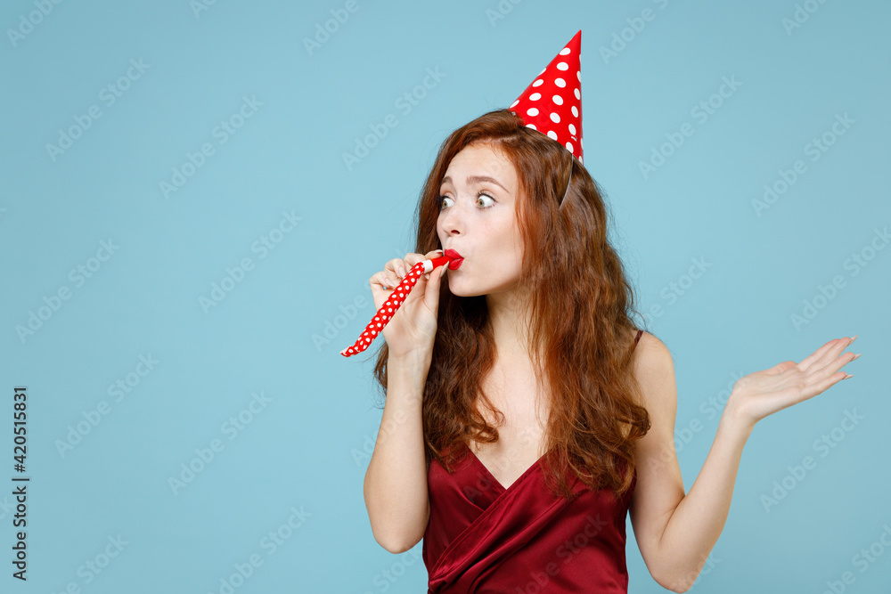 Young caucasian shocked fun happy readhead curly woman 20s wearing red party evening dress gown birthday cone hat blow pipe spread hand look aside isolated on pastel blue background studio portrait. Young caucasian shocked fun happy readhead curly woman 20s wearing red party evening dress gown birthday cone hat blow pipe spread hand look aside isolated on pastel blue background studio portrait.