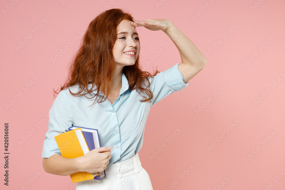 Young happy redhead student woman in blue shirt hold books, hand at forehead look far away distance isolated on pastel pink background studio portrait Education high school university college concept Young happy redhead student woman in blue shirt hold books, hand at forehead look far away distance isolated on pastel pink background studio portrait Education high school university college concept