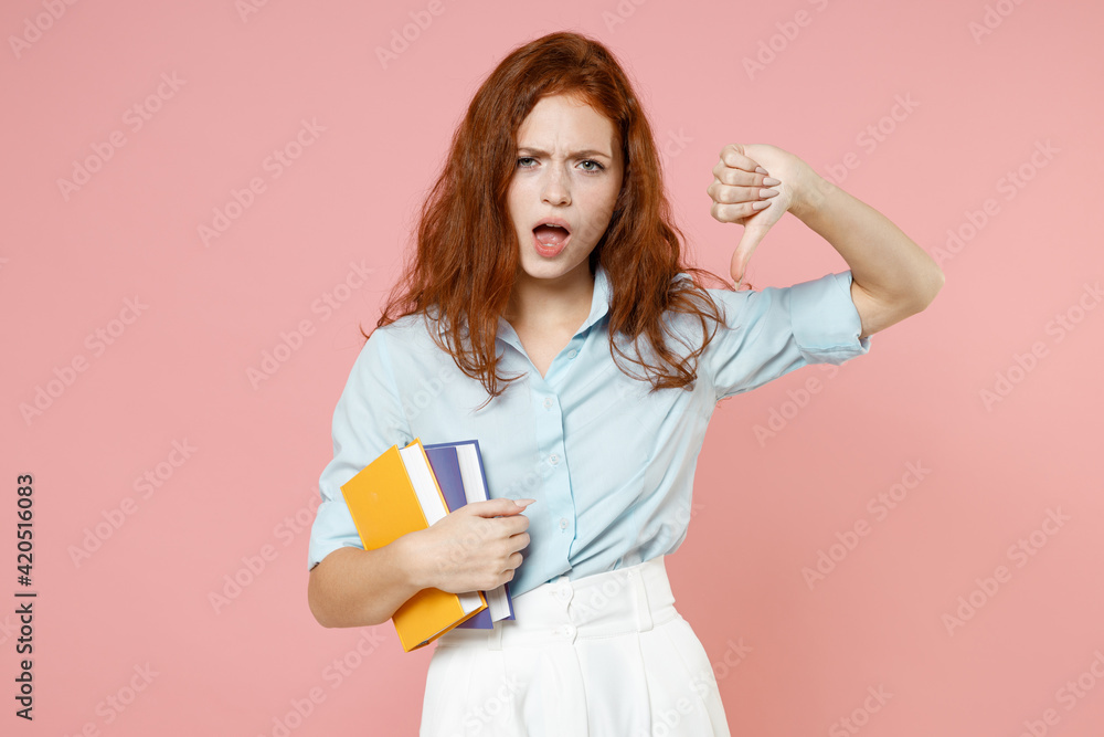 Young sad displeased redhead student woman in blue shirt hold book show thumb down dislike gesture isolated on pastel pink background studio portrait. Education high school university college concept Young sad displeased redhead student woman in blue shirt hold book show thumb down dislike gesture isolated on pastel pink background studio portrait. Education high school university college concept