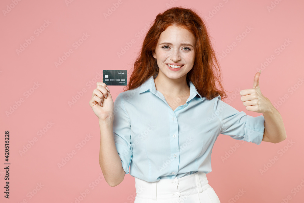 Young fun smiling rich successful student businesswoman redhead freelancer woman 20s in blue shirt holding credit bank card show thumb like gesture isolated on pastel pink background studio portrait.