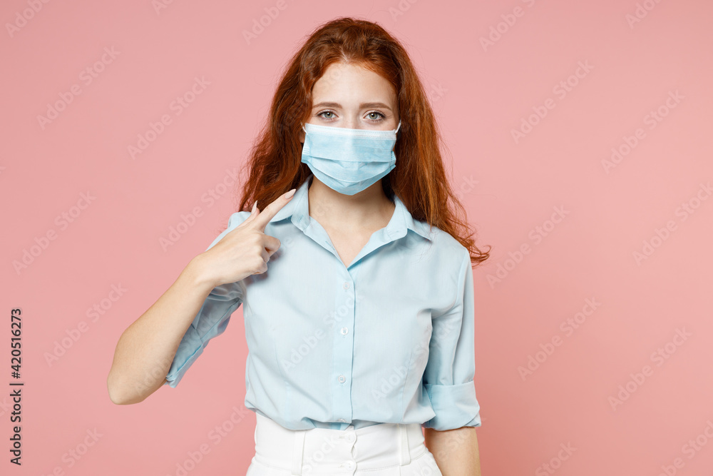 Young redhead woman 20s wear blue shirt point index finger on sterile face mask to safe from coronavirus virus covid-19 during pandemic quarantine isolated on pastel pink background studio portrait.