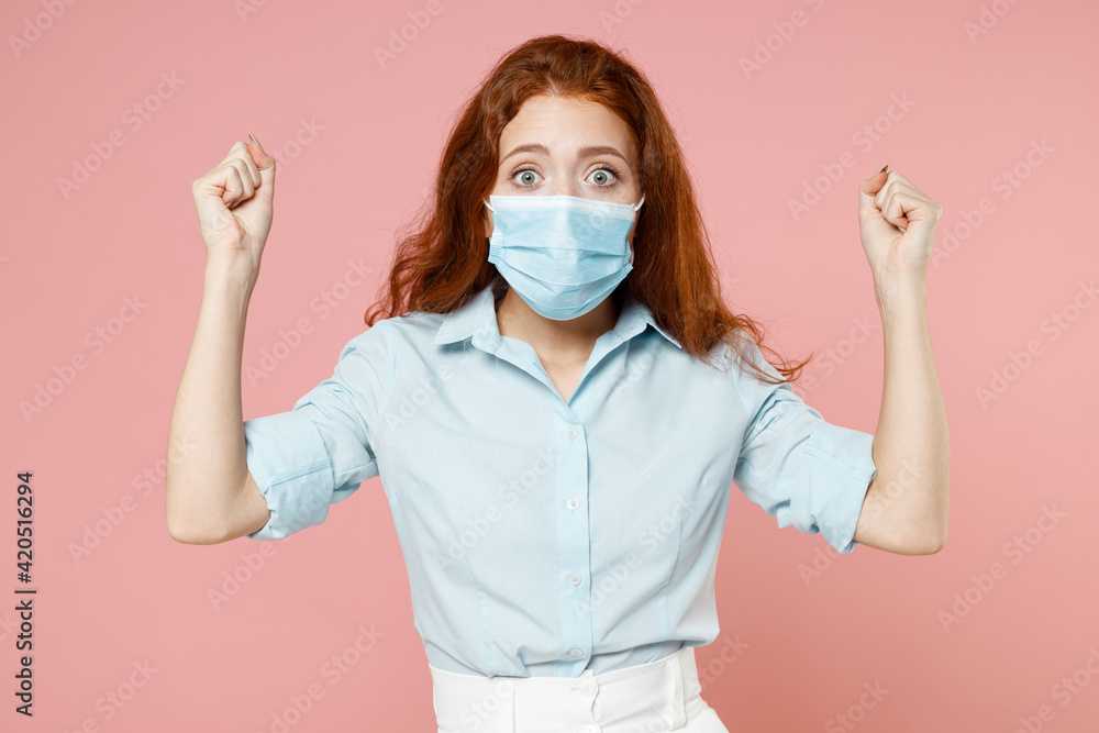 Young redhead woman 20s wear blue shirt sterile face mask from coronavirus virus covid-19 during pandemic quarantine do winnner gesture clench fist isolated on pastel pink background studio portrait. Young redhead woman 20s wear blue shirt sterile face mask from coronavirus virus covid-19 during pandemic quarantine do winnner gesture clench fist isolated on pastel pink background studio portrait.