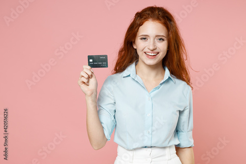 Young fun smiling rich successful student businesswoman redhead freelancer woman 20s wear blue shirt holding credit bank card looking camera isolated on pastel pink color background studio portrait