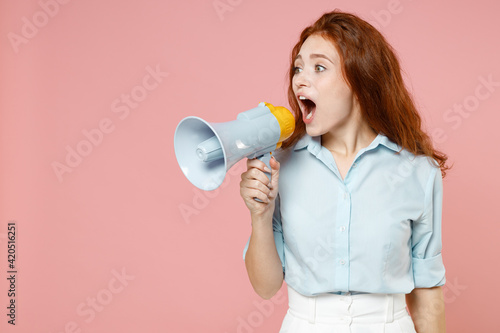 Young excited caucasain student redhead woman 20s wearing blue shirt scream loudly aside hot news shout in megaphone isolated on pastel pink color background studio portrait. People lifestyle concept.
