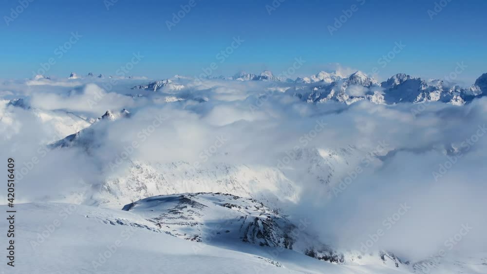 Peaks of large mountains covered with white snow and hanging soft clouds under clear blue sky in sunny winter aerial view Peaks of large mountains covered with white snow and hanging soft clouds under clear blue sky in sunny winter aerial view