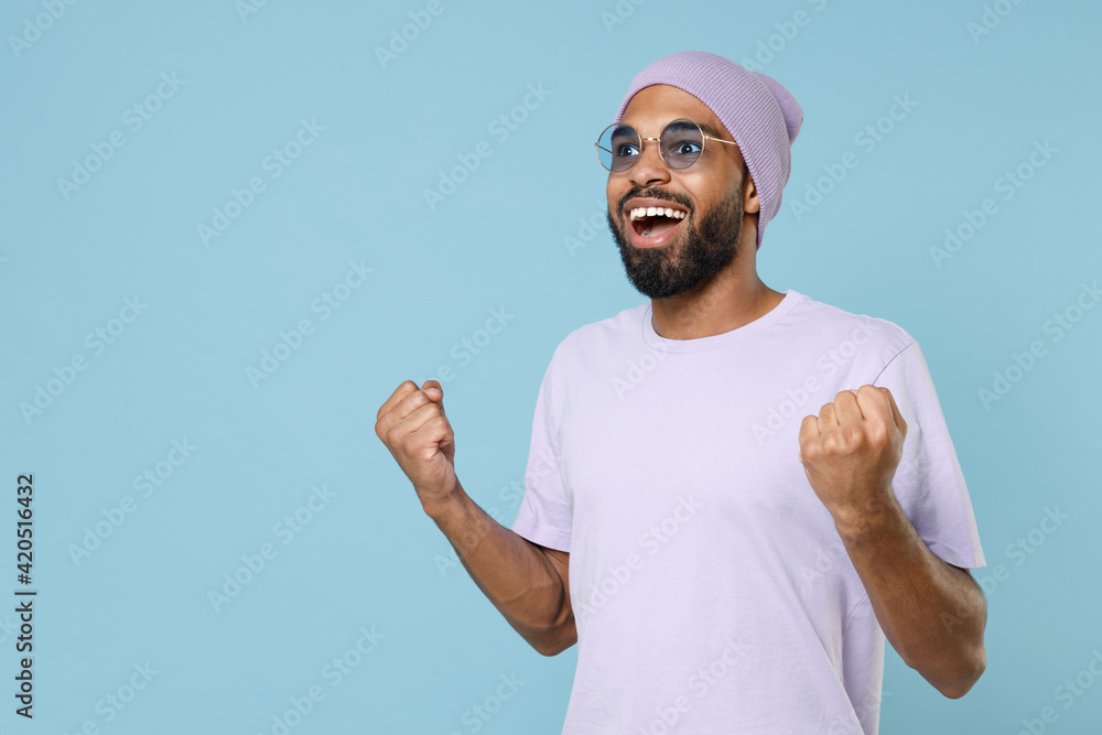 Naklejka premium Young smiling happy excited unshaven black dark-skinned african man 20s in violet t-shirt purple hat glasses do winner gesture clench fist celebrate isolated on pastel blue background studio portrait.