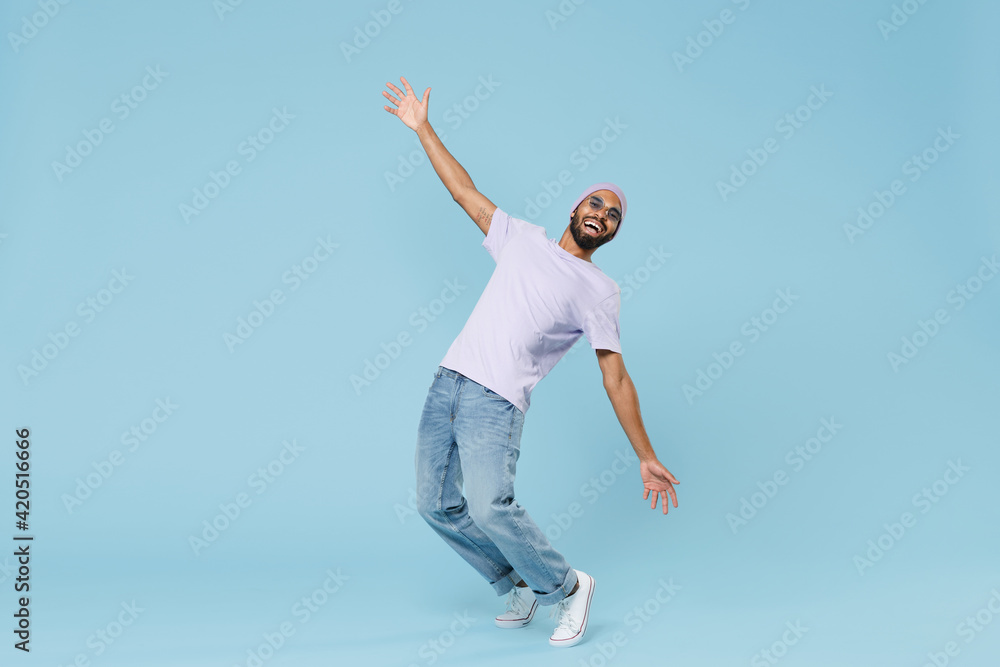 Full length young cool smiling happy funny unshaven black african man 20s wearing violet t-shirt hat glasses standing on toes leaning back dancing isolated on pastel blue background studio portrait. - obrazy, fototapety, plakaty 