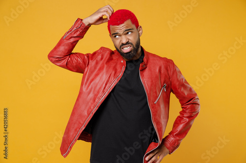 Young african dark-skinned pensive thoughtful troubled man 20s with funky trendy pink hairdo in red leather jacket look aside sctratch head isolated on yellow orange color background studio portrait photo