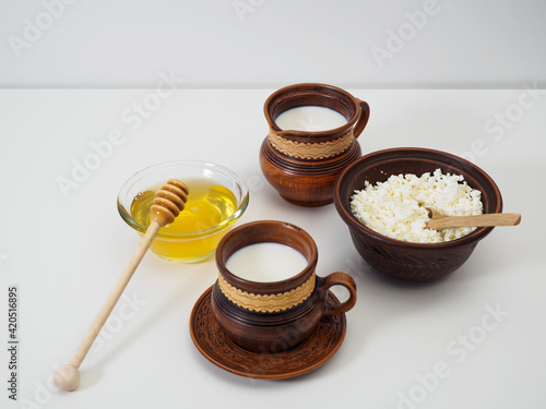 Clay jug with milk, clay cup with milk, bowl with farm cottage cheese, natural honey with honey ladle on a white table. Breakfast in earthenware. Pottery