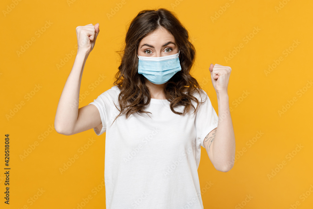 Young woman in white basic blank print design t-shirt in sterile face mask from coronavirus covid-19 pandemic quarantine do winner gesture clench fist isolated on yellow background studio portrait.