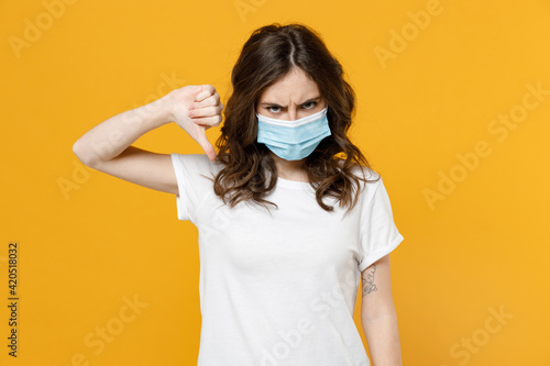 Young woman in white basic blank print design t-shirt in sterile face mask from coronavirus virus covid-19 during pandemic quarantine show thumb down dislike gesture isolated on yellow background.