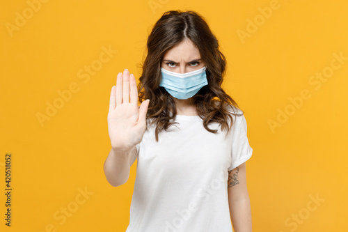 Young serious sad strict woman in white basic t-shirt in sterile face mask from coronavirus virus covid19 during pandemic quarantine do stop palm gesture refusing isolated on yellow orange background