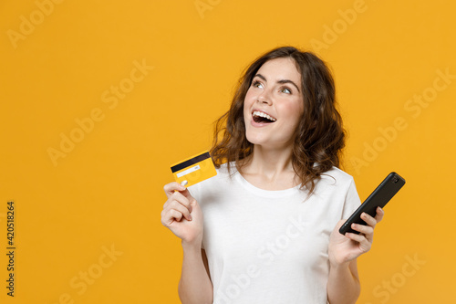 Young dreamful pensive wistful happy caucasian student woman 20s in white basic casual t-shirt look aside hold mobile cell phone credit bank card isolated on yellow orange background studio portrait.