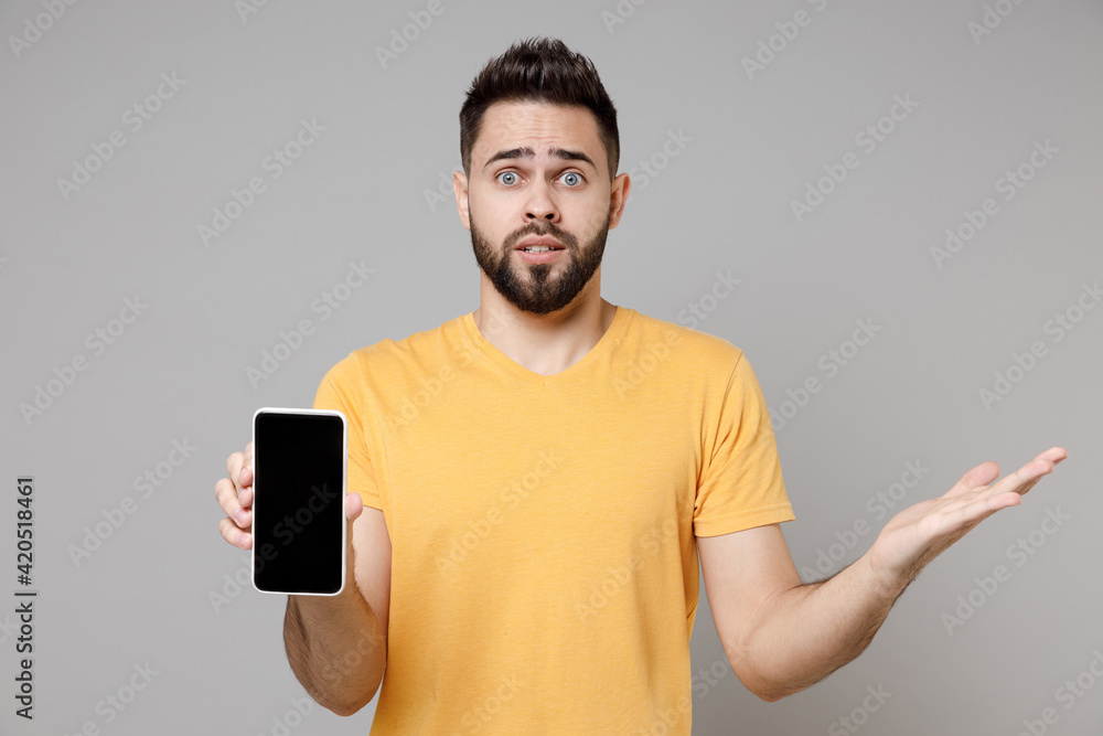 Young confused shocked bearded caucasian man 20s wear yellow basic t-shirt hold mobile cell phone, blank screen workspace area spreading hand oops gesture isolated on grey background studio portrait. Young confused shocked bearded caucasian man 20s wear yellow basic t-shirt hold mobile cell phone, blank screen workspace area spreading hand oops gesture isolated on grey background studio portrait.