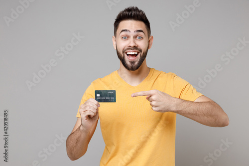 Obraz na plátně Young caucasian smiling bearded attractive rich successful happy man 20s wearing casual yellow basic t-shirt point index finger on credit bank card isolated on grey color background studio portrait