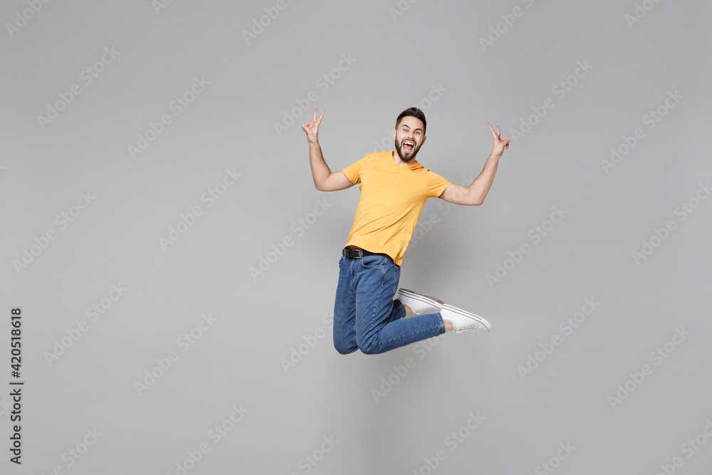 Full length of young bearded friendly successful overjoyed man 20s in yellow basic t-shirt walk show victory v-sign gesture jump high celebrating isolated on grey color background studio portrait