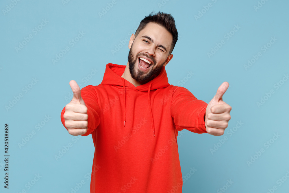 Young caucasian smiling happy bearded attractive handsome positive man 20s in casual red orange hoodie show thumbs up like gesture isolated on blue background studio portrait People lifestyle concept. Young caucasian smiling happy bearded attractive handsome positive man 20s in casual red orange hoodie show thumbs up like gesture isolated on blue background studio portrait People lifestyle concept.