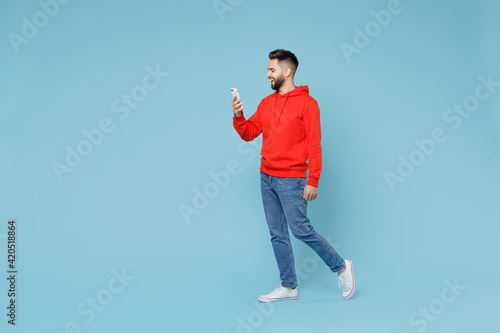 Full length side profile view of young caucasian smiling bearded attractive man 20s wear casual red orange hoodie holding mobile cell phone walking going isolated on blue background studio portrait