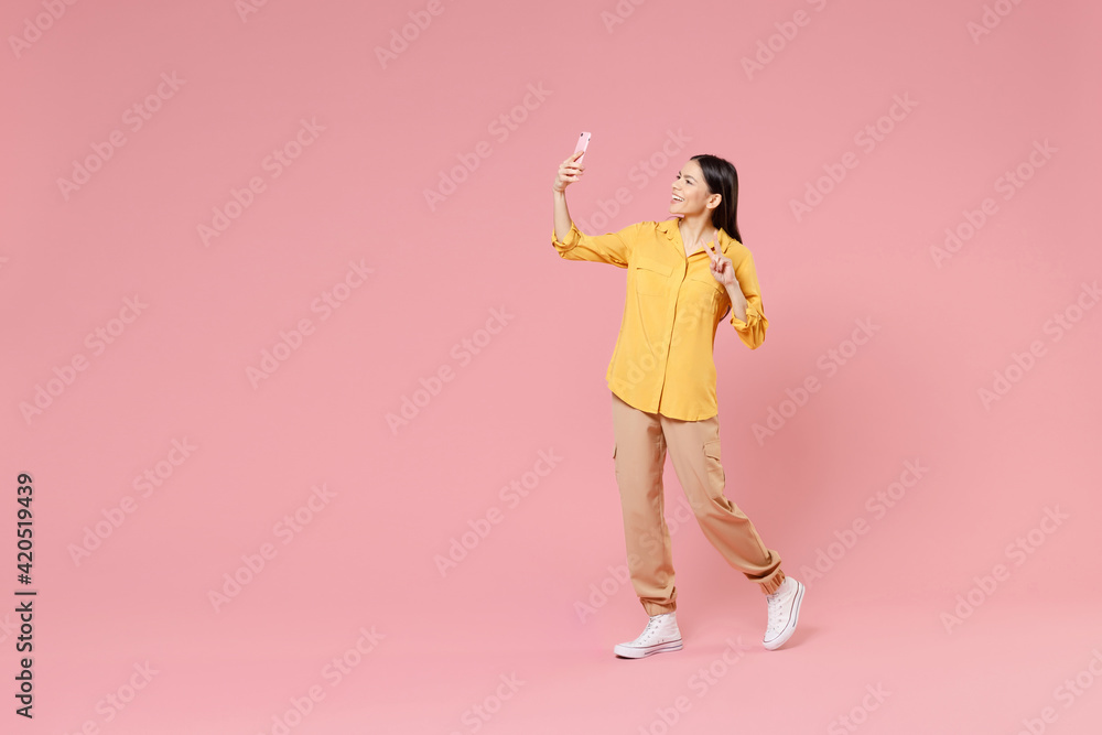 Full length side view of young smiling brunette latin woman 20s in yellow shirt doing selfie shot on mobile phone walk show victory v-sign gesture isolated on pastel pink background studio portrait. Full length side view of young smiling brunette latin woman 20s in yellow shirt doing selfie shot on mobile phone walk show victory v-sign gesture isolated on pastel pink background studio portrait.