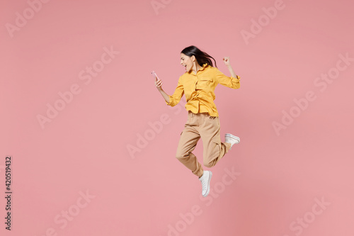 Full length side view of young brunette overjoyed latin woman 20s in yellow shirt hold mobile cell phone jump high do winner gesture clench fist isolated on pastel pink background studio portrait. © ViDi Studio