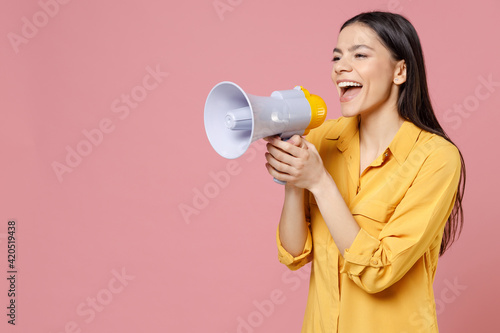 Young latin cute smiling beautiful attractive expressive excited woman 20s wearing yellow shirt screaming in megaphone shout aside hot news isolated on pastel pink color background studio portrait.