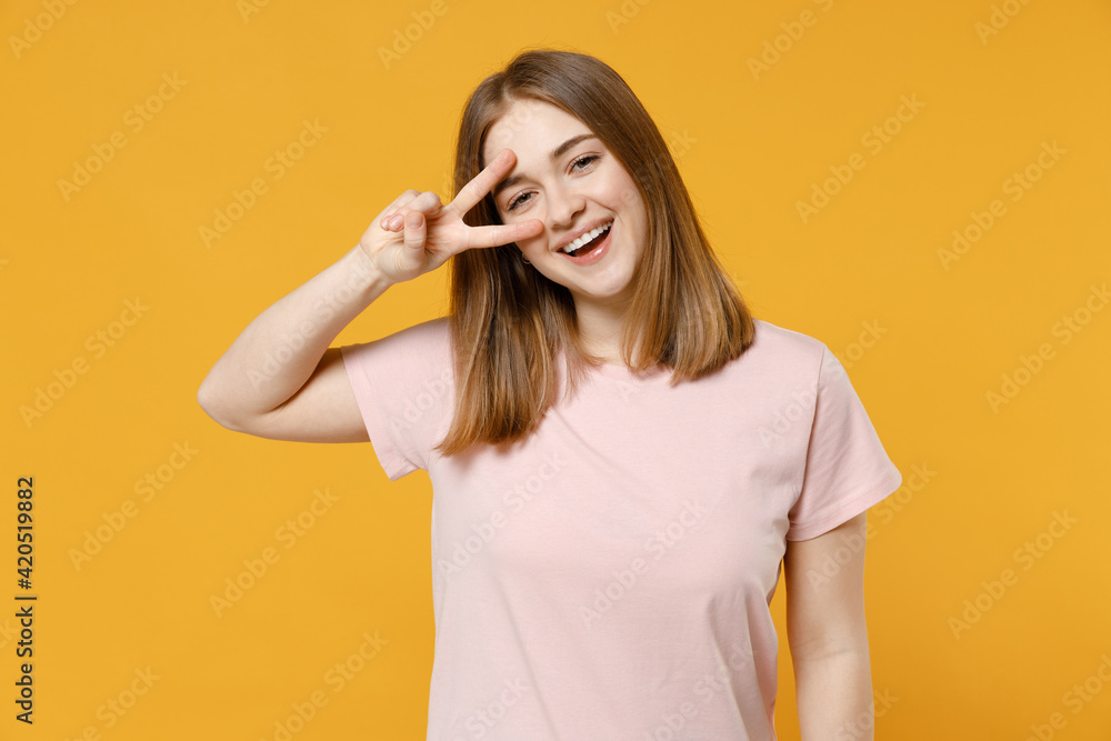 Young smiling friendly happy positive caucasian student woman 20s, nude make up in casual basic pastel pink t-shirt cover eye with victory v-sign gesture isolated on yellow background studio portrait