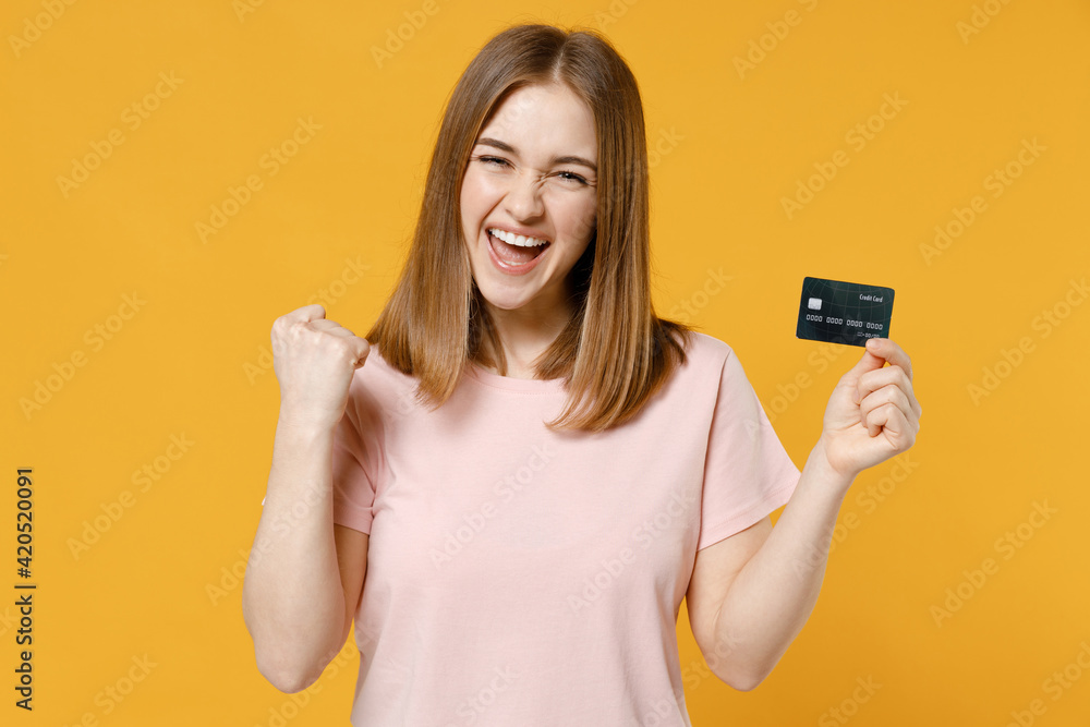 Young happy rich woman 20s in basic pastel pink t-shirt blank print design point index finger on credit bank card do winner gesture clench fist celebrate isolated on yellow background studio portrait. Young happy rich woman 20s in basic pastel pink t-shirt blank print design point index finger on credit bank card do winner gesture clench fist celebrate isolated on yellow background studio portrait.